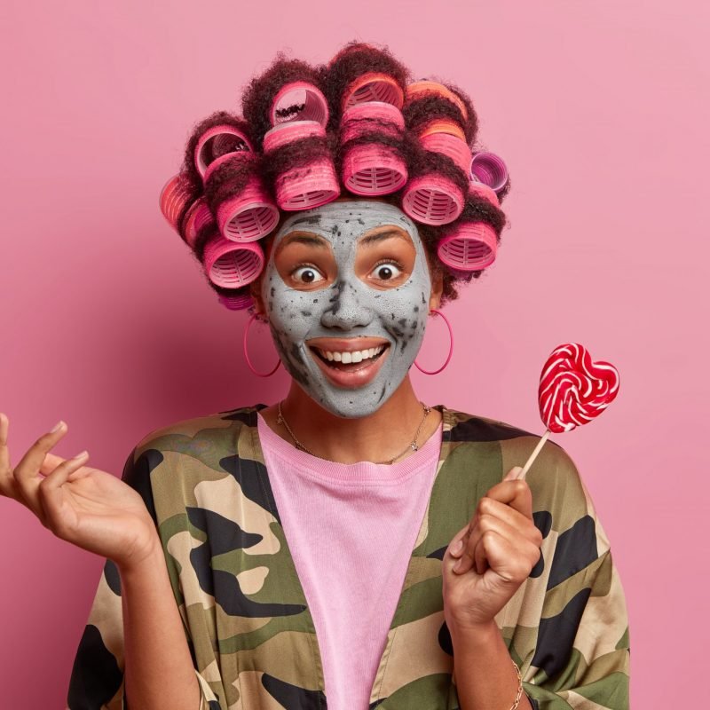 Fabulous woman looks with happy expression, raises hand and holds delicious tasty lollipop wears hair rollers, makes perfect hairstyle smiles broadly dressed in khaki dressing gown beauty mask on face