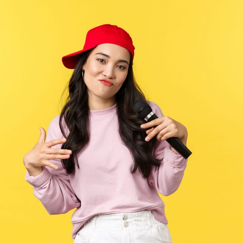 People emotions, lifestyle leisure and beauty concept. Stylish and cool young girl rapper in red cap, singing song and gesturing, performing with microphone, standing yellow background.